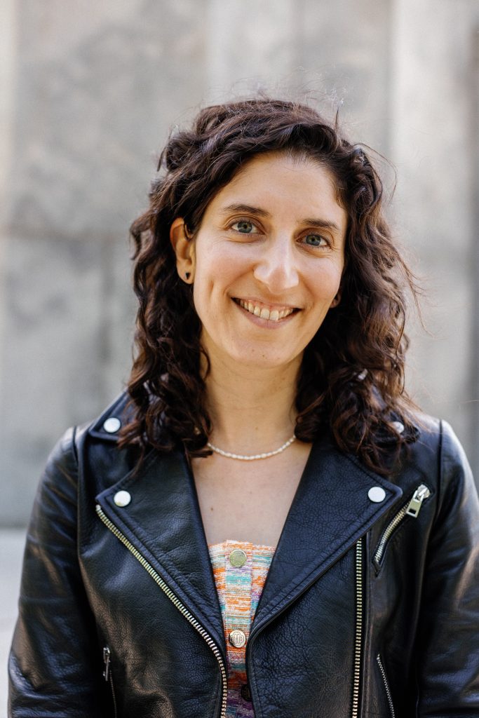 This photo shows Sara, a white woman with brown wavy medium length hair, wearing a black leather jacket.